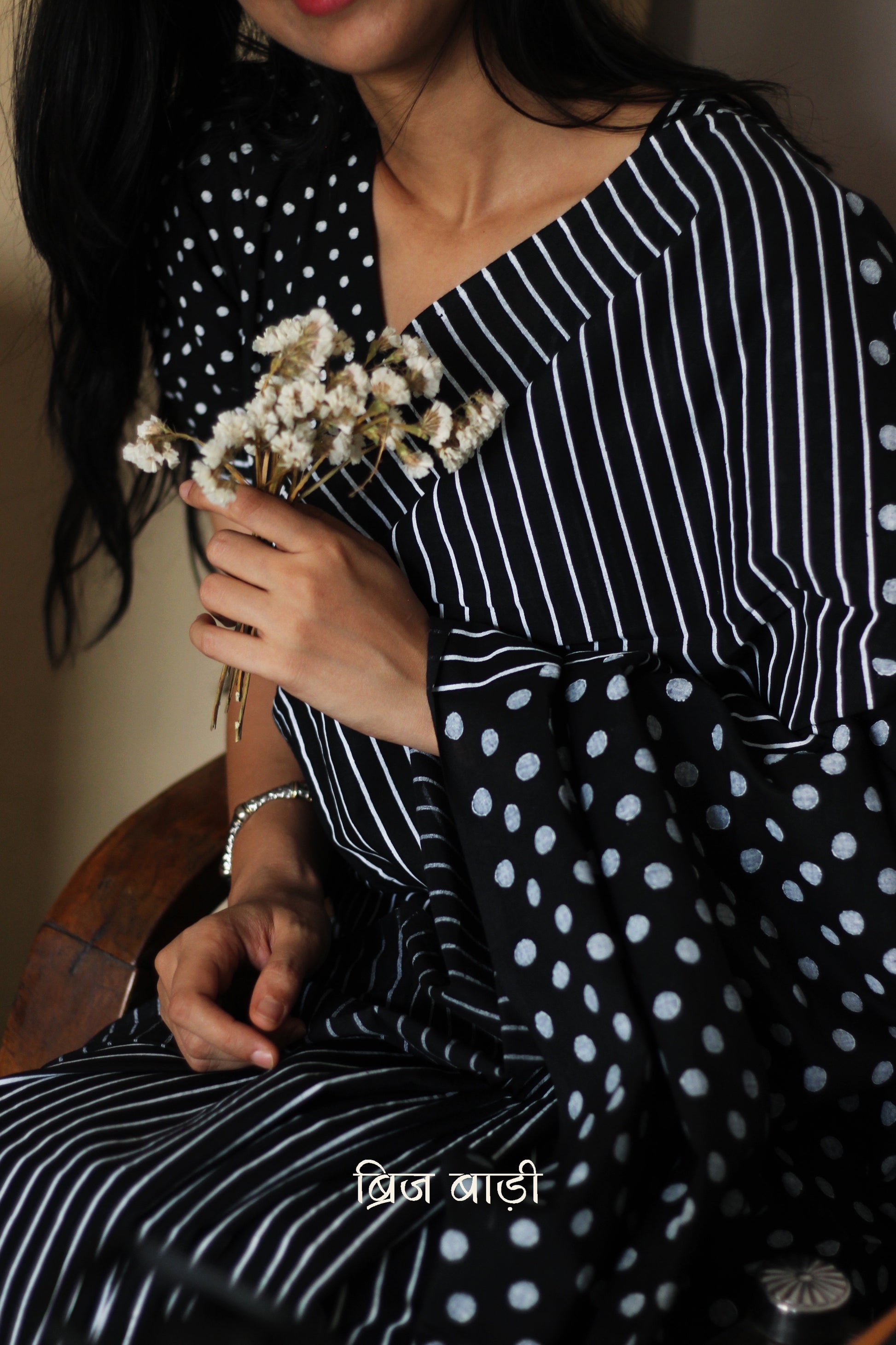 Black soft cotton saree by Brij Bari, featuring a bold and striking hand block printed pattern of stripes and polka dots for a modern, playful twist. Handcrafted from breathable, light-weight cotton, the saree drapes effortlessly—ideal for both everyday wear and special occasions. Finished with handcrafted red thread tassels on the pallu, adding an artisanal touch of charm. It comes with a free hand block printed blouse piece that complements the print and palette. 