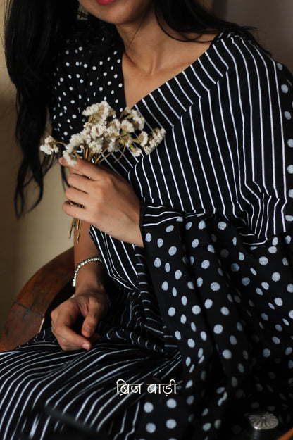 Black soft cotton saree by Brij Bari, featuring a bold and striking hand block printed pattern of stripes and polka dots for a modern, playful twist. Handcrafted from breathable, light-weight cotton, the saree drapes effortlessly—ideal for both everyday wear and special occasions. Finished with handcrafted red thread tassels on the pallu, adding an artisanal touch of charm. It comes with a free hand block printed blouse piece that complements the print and palette. 