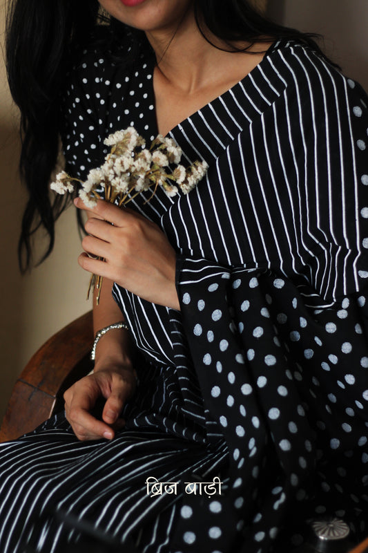Black soft cotton saree by Brij Bari, featuring a bold and striking hand block printed pattern of stripes and polka dots for a modern, playful twist. Handcrafted from breathable, light-weight cotton, the saree drapes effortlessly—ideal for both everyday wear and special occasions. Finished with handcrafted red thread tassels on the pallu, adding an artisanal touch of charm. It comes with a free hand block printed blouse piece that complements the print and palette. 