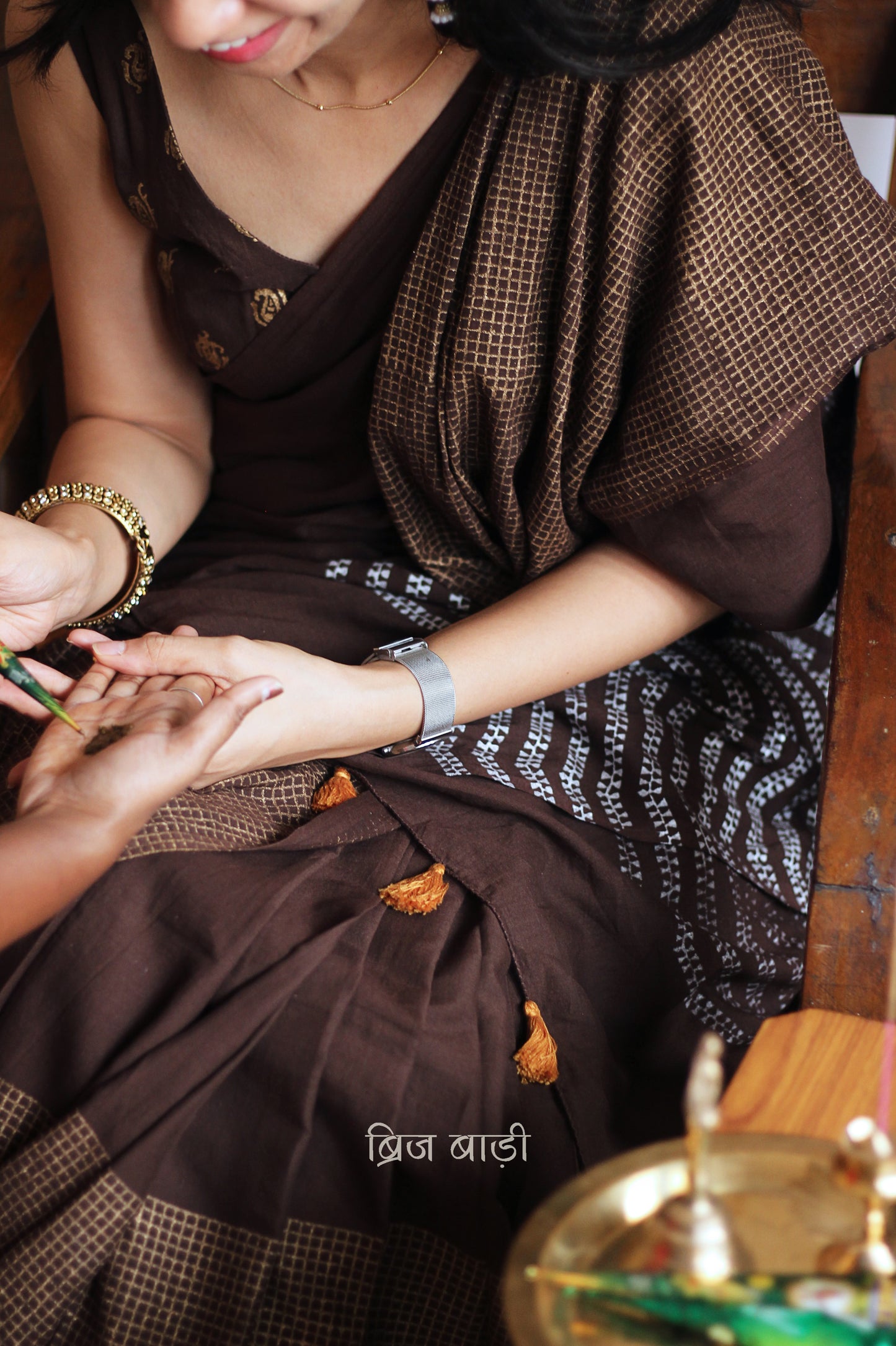 Earthy brown light-weight cotton saree by Brij Bari, featuring intricate golden hand block printed check patterns on the skirt and silver geometric designs on the pallu. Handcrafted mustard thread tassels add a warm, artisanal touch to the pallu. Includes a free hand block printed blouse piece that complements the saree’s festive and versatile appeal.