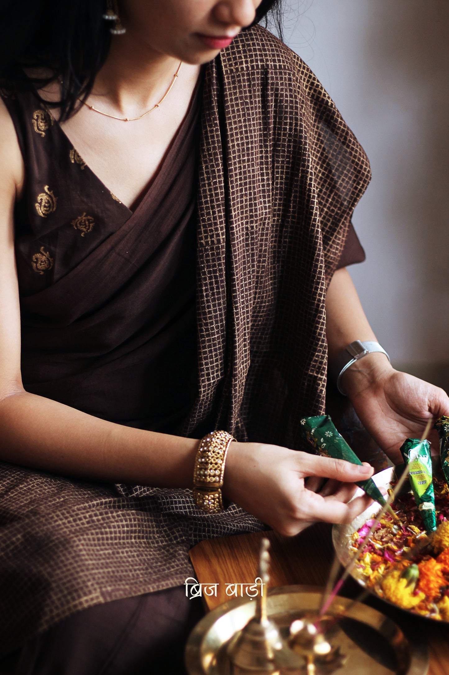 Earthy brown light-weight cotton saree by Brij Bari, featuring intricate golden hand block printed check patterns on the skirt and silver geometric designs on the pallu. Handcrafted mustard thread tassels add a warm, artisanal touch to the pallu. Includes a free hand block printed blouse piece that complements the saree’s festive and versatile appeal.