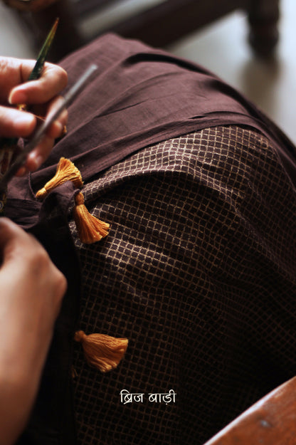 Earthy brown light-weight cotton saree by Brij Bari, featuring intricate golden hand block printed check patterns on the skirt and silver geometric designs on the pallu. Handcrafted mustard thread tassels add a warm, artisanal touch to the pallu. Includes a free hand block printed blouse piece that complements the saree’s festive and versatile appeal.