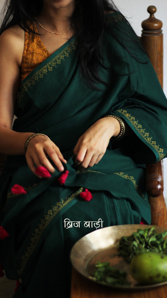 Deep green soft and light-weight cotton saree by Brij Bari, featuring a delicate hand block printed border in vibrant ochre yellow that contrasts beautifully with the rich green. The saree is finished with pink handmade thread tassels on the pallu, adding a touch of traditional charm. It comes with a free matching block printed blouse piece, completing the handcrafted look. Perfect for everyday wear, casual outings, or semi-formal occasions, with free shipping pan India.