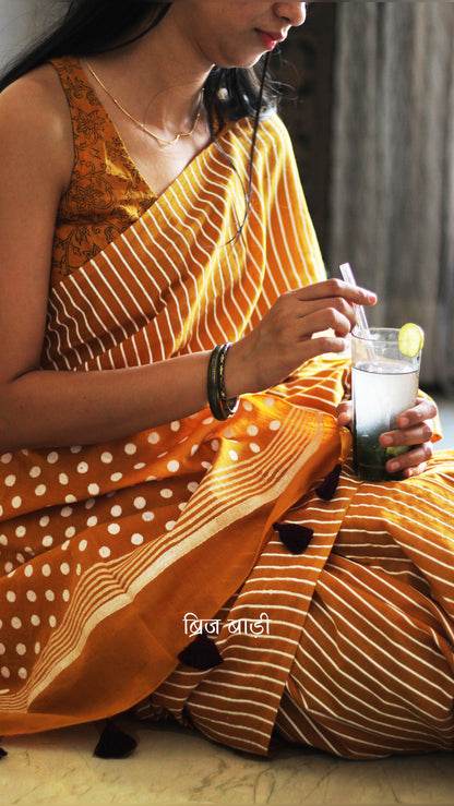 Ochre Yellow soft cotton saree by Brij Bari, featuring a bold and striking hand block printed pattern of stripes and polka dots for a modern, playful twist. Handcrafted from breathable, light-weight cotton, the saree drapes effortlessly—ideal for both everyday wear and special occasions. Finished with handcrafted purple thread tassels on the pallu, adding an artisanal touch of charm. It comes with a free hand block printed blouse piece that complements the print and palette. 