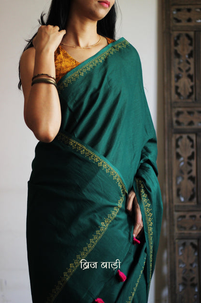 Deep green soft and light-weight cotton saree by Brij Bari, featuring a delicate hand block printed border in vibrant ochre yellow that contrasts beautifully with the rich green. The saree is finished with pink handmade thread tassels on the pallu, adding a touch of traditional charm. It comes with a free matching block printed blouse piece, completing the handcrafted look. Perfect for everyday wear, casual outings, or semi-formal occasions, with free shipping pan India.