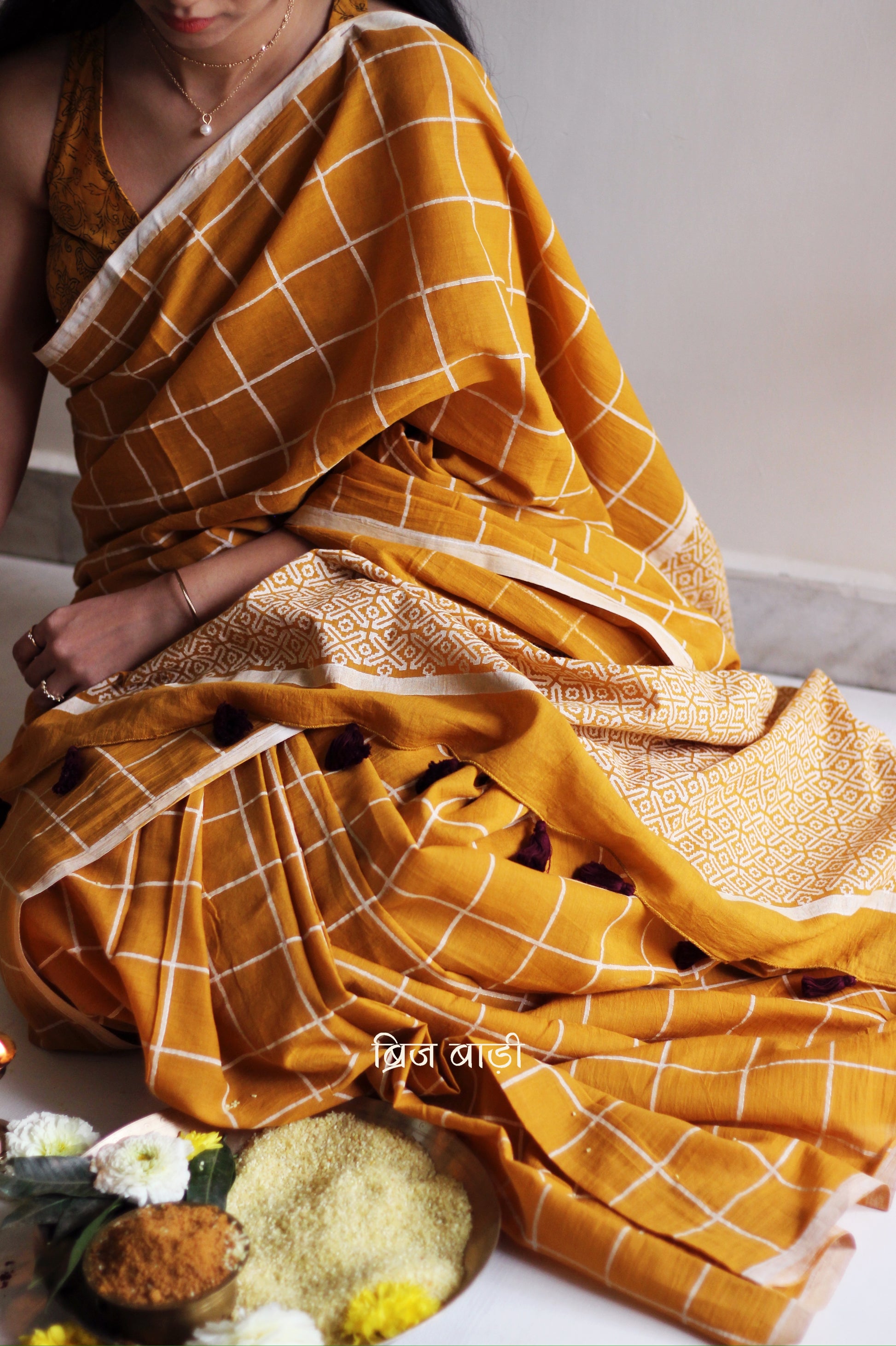 Yellow soft and light-weight cotton saree by Brij Bari, hand block printed in white with a classic check pattern on the body and a striking geometrical pattern on the pallu. Detailed with pink handmade thread tassels that add a charming, handcrafted touch. This saree comes with a free matching block printed blouse piece, offering a complete, artisanal look. A versatile, breathable saree perfect for everyday wear, casual outings, or semi-formal occasions, with free shipping pan India. Available in 2 colours