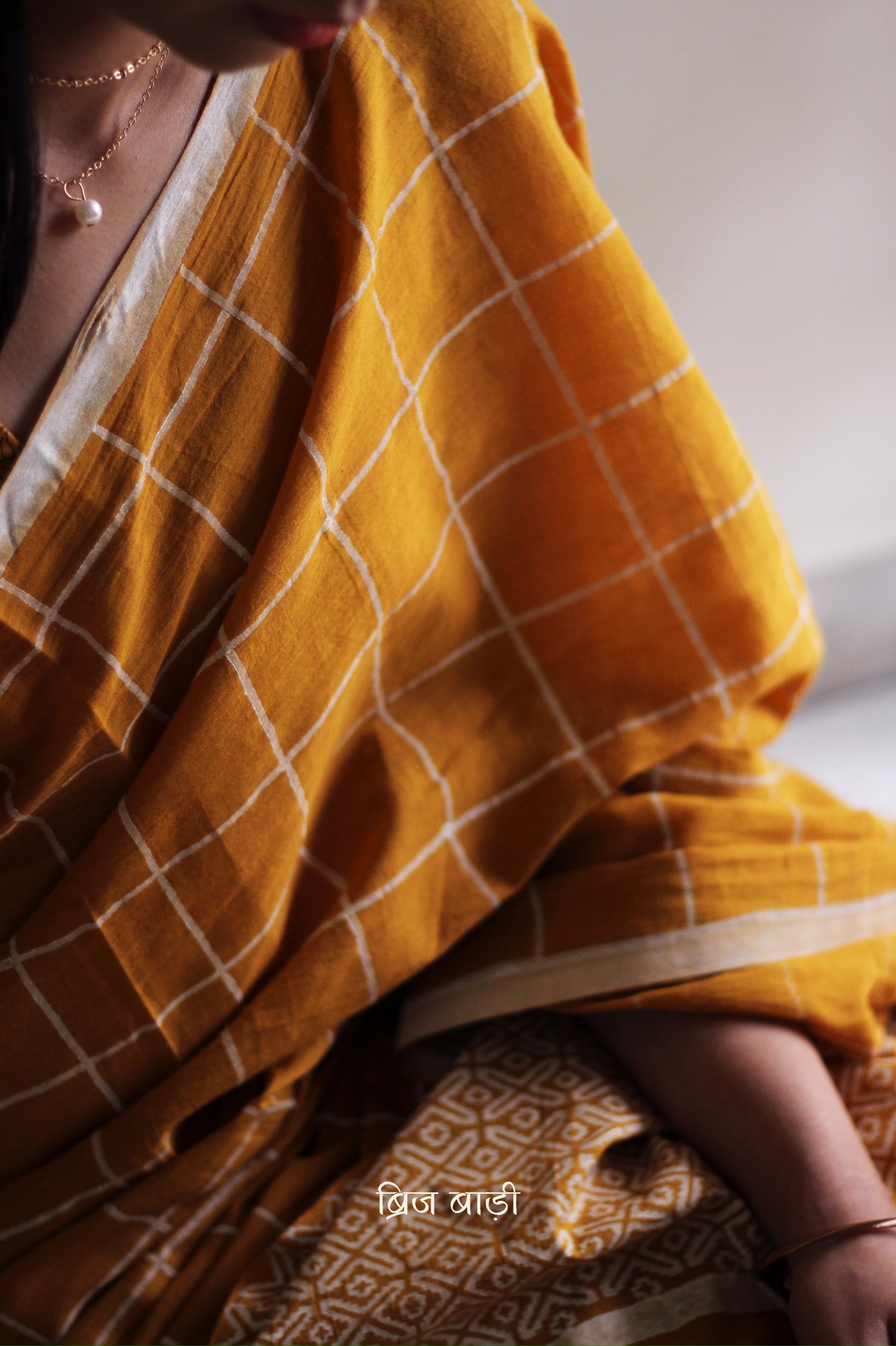 Yellow soft and light-weight cotton saree by Brij Bari, hand block printed in white with a classic check pattern on the body and a striking geometrical pattern on the pallu. Detailed with pink handmade thread tassels that add a charming, handcrafted touch. This saree comes with a free matching block printed blouse piece, offering a complete, artisanal look. A versatile, breathable saree perfect for everyday wear, casual outings, or semi-formal occasions, with free shipping pan India. Available in 2 colours