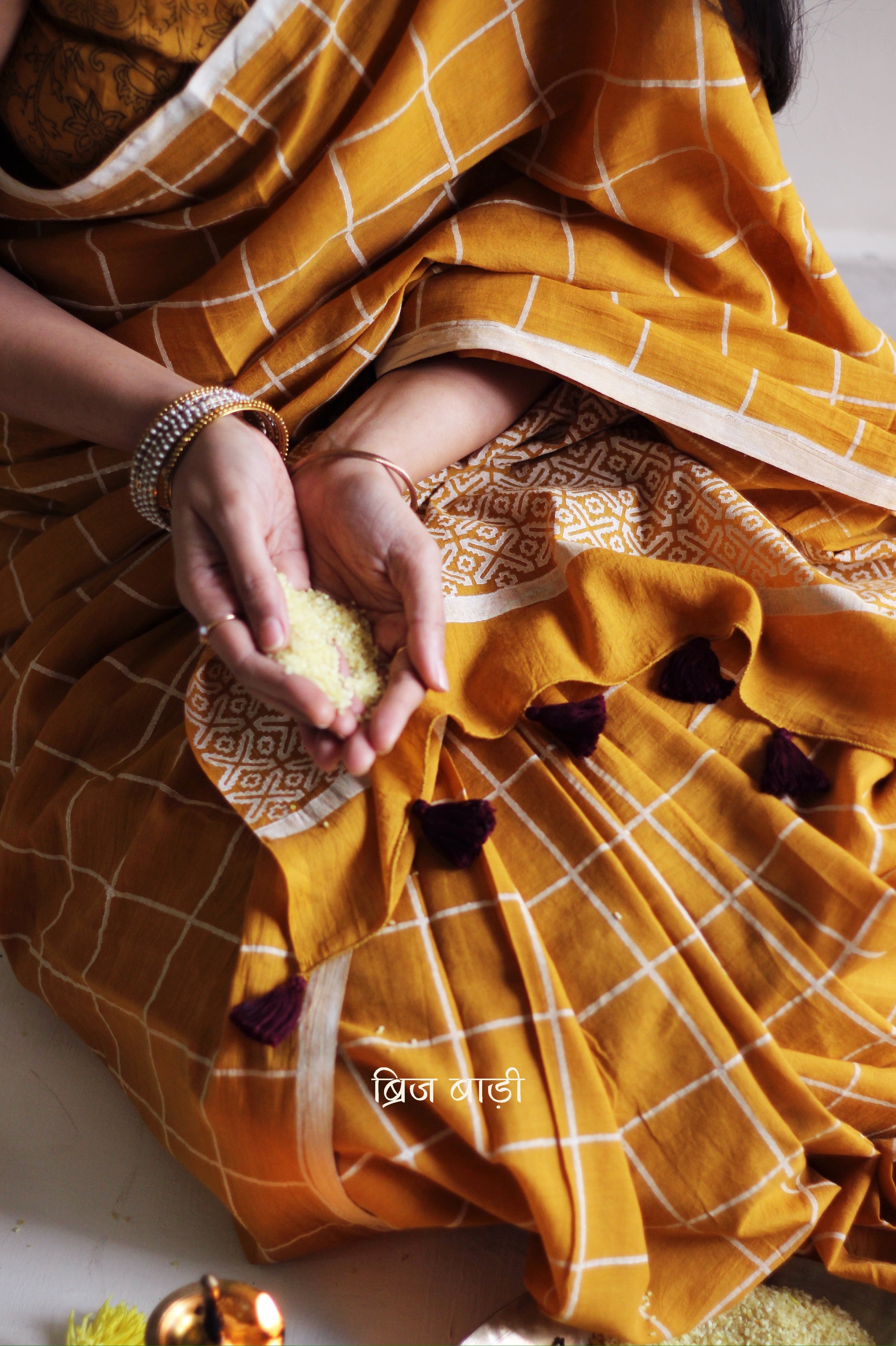 Yellow soft and light-weight cotton saree by Brij Bari, hand block printed in white with a classic check pattern on the body and a striking geometrical pattern on the pallu. Detailed with pink handmade thread tassels that add a charming, handcrafted touch. This saree comes with a free matching block printed blouse piece, offering a complete, artisanal look. A versatile, breathable saree perfect for everyday wear, casual outings, or semi-formal occasions, with free shipping pan India. Available in 2 colours