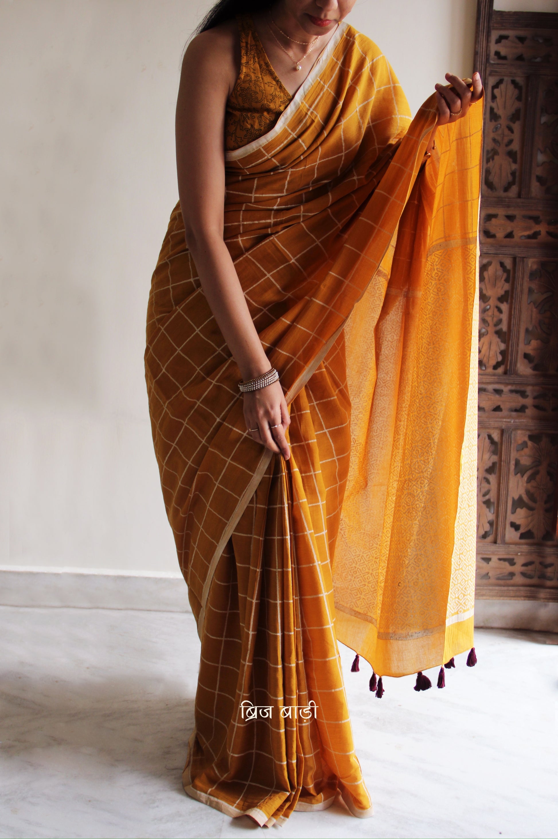 Yellow soft and light-weight cotton saree by Brij Bari, hand block printed in white with a classic check pattern on the body and a striking geometrical pattern on the pallu. Detailed with pink handmade thread tassels that add a charming, handcrafted touch. This saree comes with a free matching block printed blouse piece, offering a complete, artisanal look. A versatile, breathable saree perfect for everyday wear, casual outings, or semi-formal occasions, with free shipping pan India. Available in 2 colours