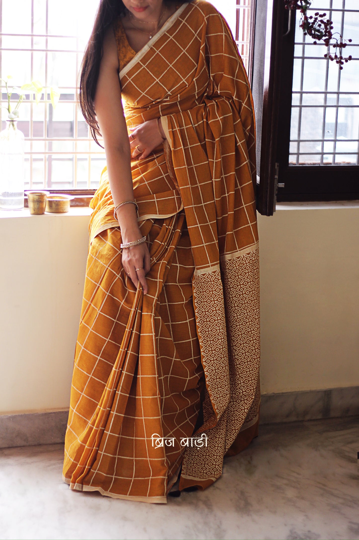 Yellow soft and light-weight cotton saree by Brij Bari, hand block printed in white with a classic check pattern on the body and a striking geometrical pattern on the pallu. Detailed with pink handmade thread tassels that add a charming, handcrafted touch. This saree comes with a free matching block printed blouse piece, offering a complete, artisanal look. A versatile, breathable saree perfect for everyday wear, casual outings, or semi-formal occasions, with free shipping pan India. Available in 2 colours