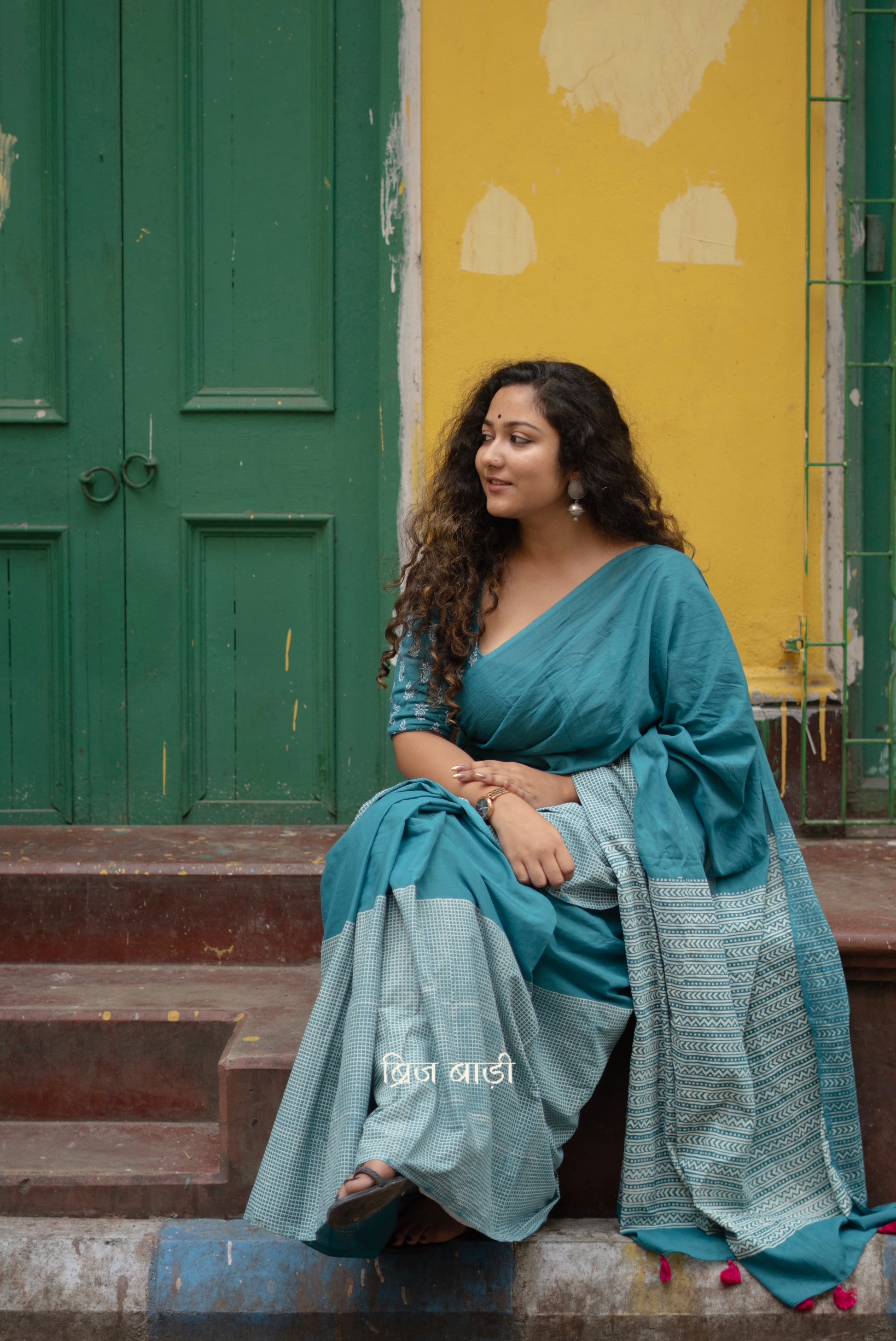 Teal blue soft and lightweight cotton saree by Brij Bari, hand block printed with a classic check pattern on the skirt and a geometric design in white on the pallu. Finished with handmade orange thread tassels on the pallu. Comes with a complimentary block printed cotton blouse piece. A versatile design that adds a pop of colour to your everyday wardrobe while offering breathable comfort and effortless style. Free shipping pan India.