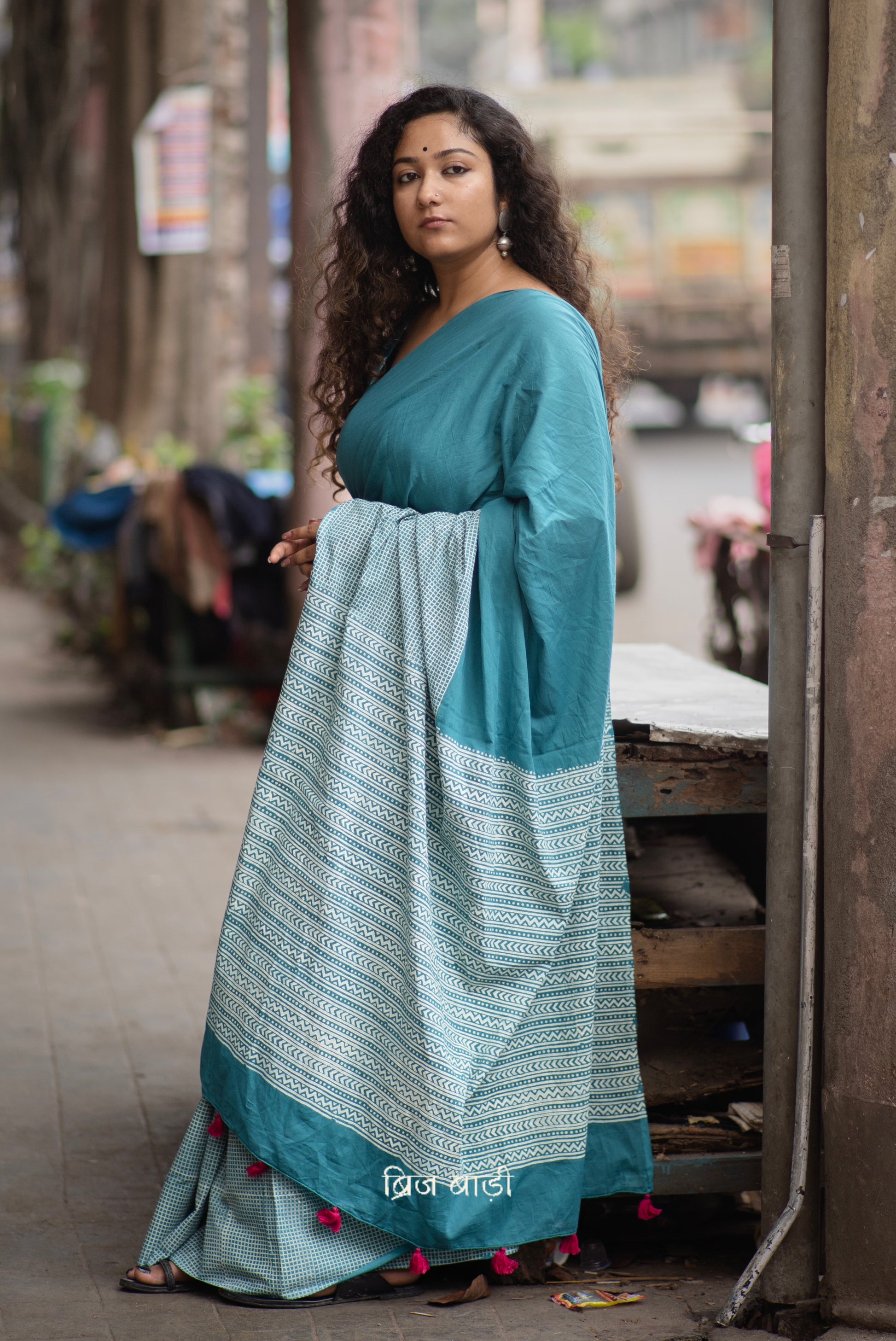 Teal blue soft and lightweight cotton saree by Brij Bari, hand block printed with a classic check pattern on the skirt and a geometric design in white on the pallu. Finished with handmade orange thread tassels on the pallu. Comes with a complimentary block printed cotton blouse piece. A versatile design that adds a pop of colour to your everyday wardrobe while offering breathable comfort and effortless style. Free shipping pan India.