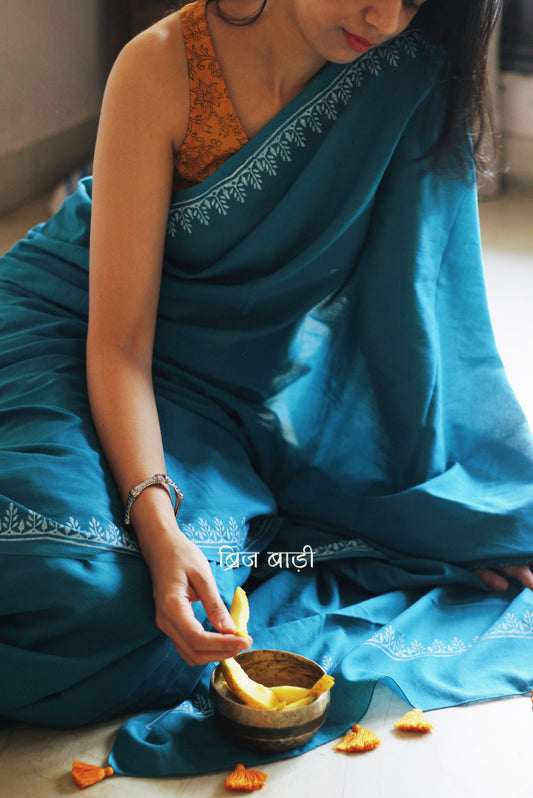 Teal blue soft and light-weight cotton saree by Brij Bari, designed for all-day comfort and effortless elegance. Detailed with a delicate hand block printed border in crisp white, creating a refined contrast. The saree is finished with orange handmade thread tassels on the pallu, adding a subtle pop of colour. Comes with a free matching block printed blouse piece. Ideal for everyday wear, office, casual outings, or light festive occasions, with free shipping pan India.