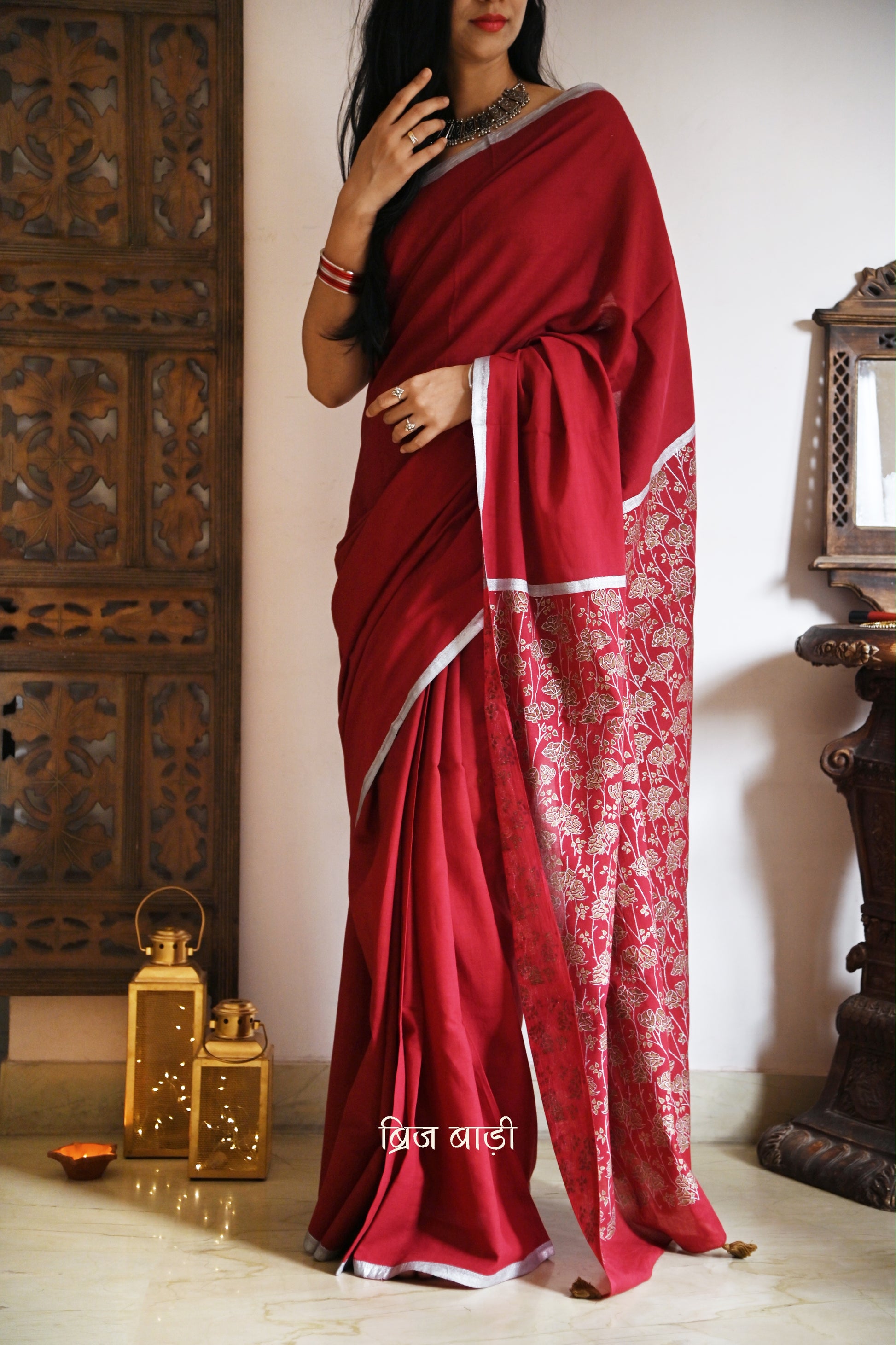 Deep red light-weight cotton saree by Brij Bari, featuring an intricately hand block printed floral pattern in gold and silver on the pallu. The saree is designed with a minimalist border for a timeless appeal, and the pallu is adorned with handcrafted olive green thread tassels that add a unique, earthy contrast. A versatile and comfortable pick for the festive season, it comes with a free matching hand block printed blouse piece. Thoughtfully crafted for elegance, ease, and tradition.

