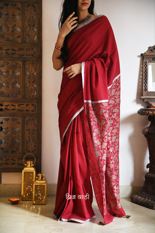 Deep red light-weight cotton saree by Brij Bari, featuring an intricately hand block printed floral pattern in gold and silver on the pallu. The saree is designed with a minimalist border for a timeless appeal, and the pallu is adorned with handcrafted olive green thread tassels that add a unique, earthy contrast. A versatile and comfortable pick for the festive season, it comes with a free matching hand block printed blouse piece. Thoughtfully crafted for elegance, ease, and tradition.

