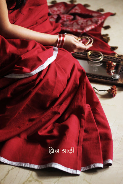 Deep red light-weight cotton saree by Brij Bari, featuring an intricately hand block printed floral pattern in gold and silver on the pallu. The saree is designed with a minimalist border for a timeless appeal, and the pallu is adorned with handcrafted olive green thread tassels that add a unique, earthy contrast. A versatile and comfortable pick for the festive season, it comes with a free matching hand block printed blouse piece. Thoughtfully crafted for elegance, ease, and tradition.

