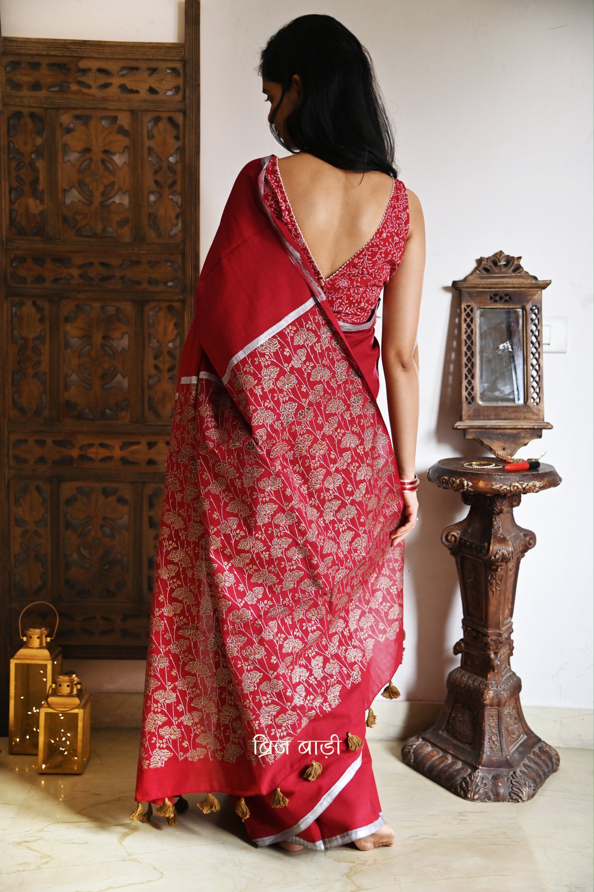 Deep red light-weight cotton saree by Brij Bari, featuring an intricately hand block printed floral pattern in gold and silver on the pallu. The saree is designed with a minimalist border for a timeless appeal, and the pallu is adorned with handcrafted olive green thread tassels that add a unique, earthy contrast. A versatile and comfortable pick for the festive season, it comes with a free matching hand block printed blouse piece. Thoughtfully crafted for elegance, ease, and tradition.

