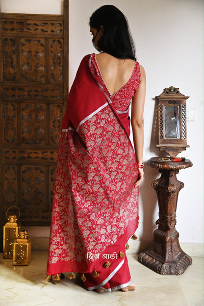 Deep red light-weight cotton saree by Brij Bari, featuring an intricately hand block printed floral pattern in gold and silver on the pallu. The saree is designed with a minimalist border for a timeless appeal, and the pallu is adorned with handcrafted olive green thread tassels that add a unique, earthy contrast. A versatile and comfortable pick for the festive season, it comes with a free matching hand block printed blouse piece. Thoughtfully crafted for elegance, ease, and tradition.

