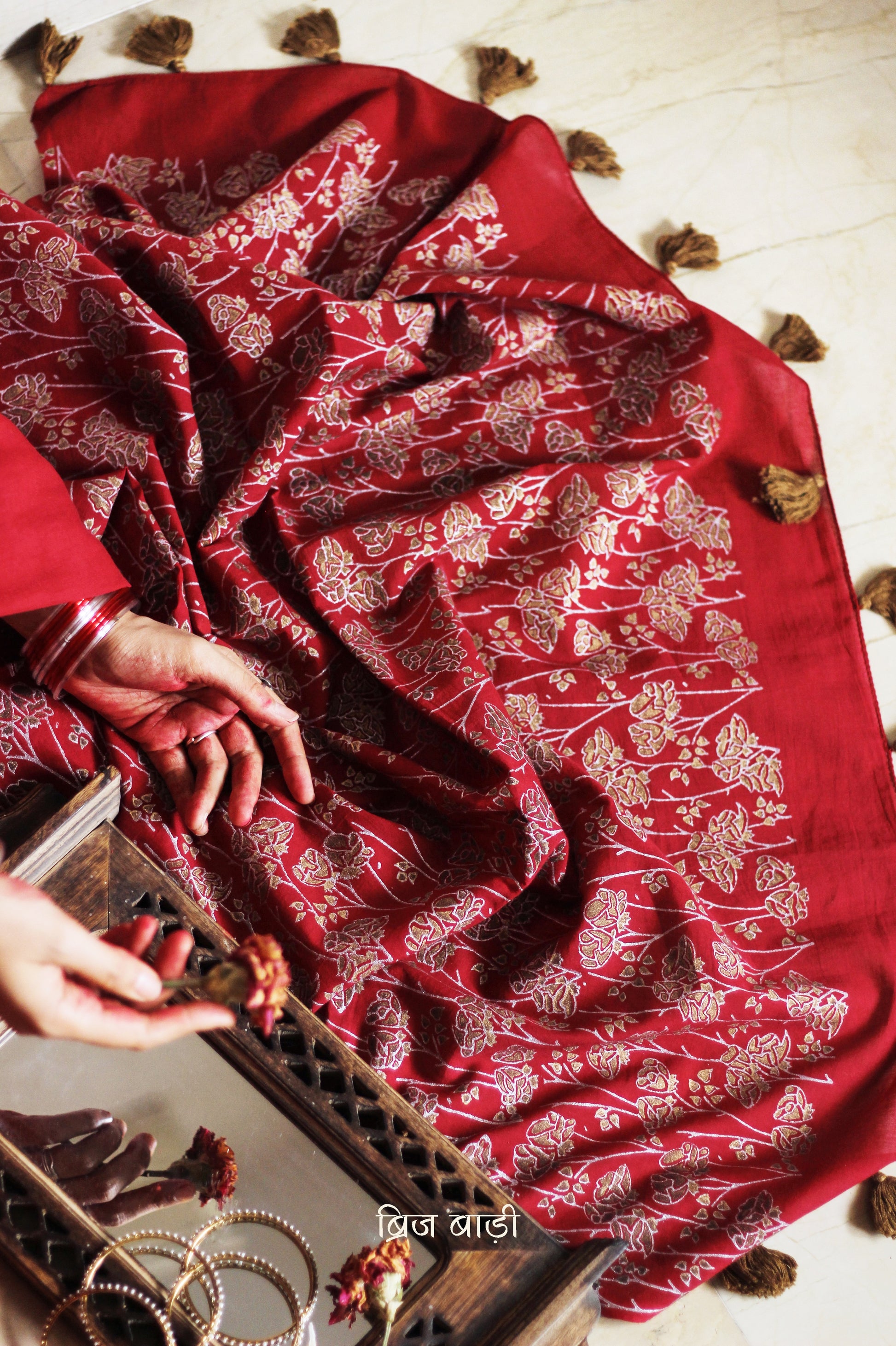 Deep red light-weight cotton saree by Brij Bari, featuring an intricately hand block printed floral pattern in gold and silver on the pallu. The saree is designed with a minimalist border for a timeless appeal, and the pallu is adorned with handcrafted olive green thread tassels that add a unique, earthy contrast. A versatile and comfortable pick for the festive season, it comes with a free matching hand block printed blouse piece. Thoughtfully crafted for elegance, ease, and tradition.

