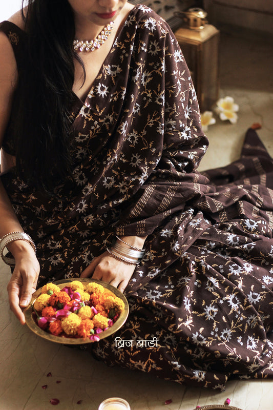 Brown soft cotton saree by Brij Bari, adorned with an abstract floral hand block print in elegant shades of gold and silver, lending it a refined festive charm. Crafted from breathable, light-weight cotton, the saree is accented with handcrafted mustard thread tassels on the pallu for a warm, artisanal finish. It comes with a free hand block printed blouse piece that complements the overall design. Ideal for festive occasions, this saree blends traditional techniques with modern aesthetics