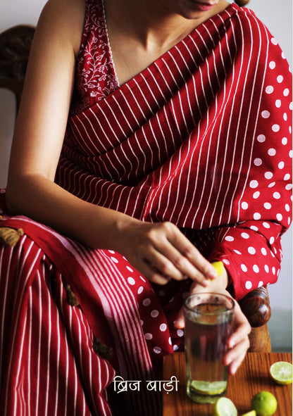 Deep Red soft cotton saree by Brij Bari, featuring a bold and striking hand block printed pattern of stripes and polka dots for a modern, playful twist. Crafted from breathable, light-weight cotton, the saree drapes effortlessly—ideal for both everyday wear and special occasions. Finished with handcrafted olive green thread tassels on the pallu, adding an artisanal touch of charm. It comes with a free hand block printed blouse piece that complements the print and palette. 