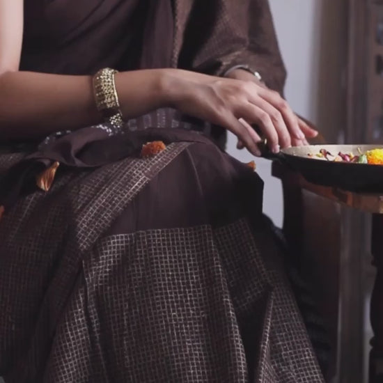 Earthy brown light-weight cotton saree by Brij Bari, featuring intricate golden hand block printed check patterns on the skirt and silver geometric designs on the pallu. Handcrafted mustard thread tassels add a warm, artisanal touch to the pallu. Includes a free hand block printed blouse piece that complements the saree’s festive and versatile appeal.
