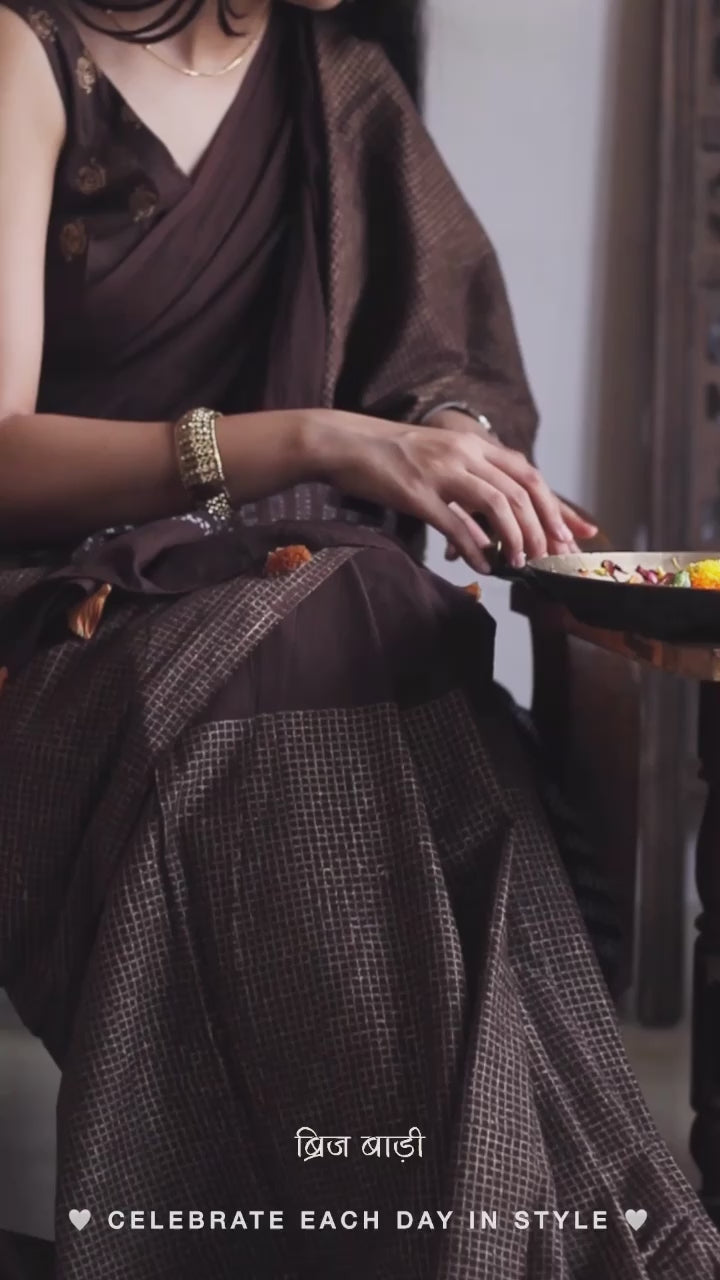 Earthy brown light-weight cotton saree by Brij Bari, featuring intricate golden hand block printed check patterns on the skirt and silver geometric designs on the pallu. Handcrafted mustard thread tassels add a warm, artisanal touch to the pallu. Includes a free hand block printed blouse piece that complements the saree’s festive and versatile appeal.