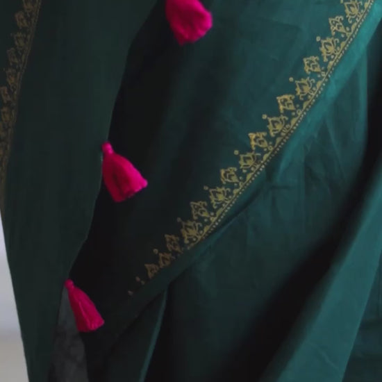 Deep green soft and light-weight cotton saree by Brij Bari, featuring a delicate hand block printed border in vibrant ochre yellow that contrasts beautifully with the rich green. The saree is finished with pink handmade thread tassels on the pallu, adding a touch of traditional charm. It comes with a free matching block printed blouse piece, completing the handcrafted look. Perfect for everyday wear, casual outings, or semi-formal occasions, with free shipping pan India.