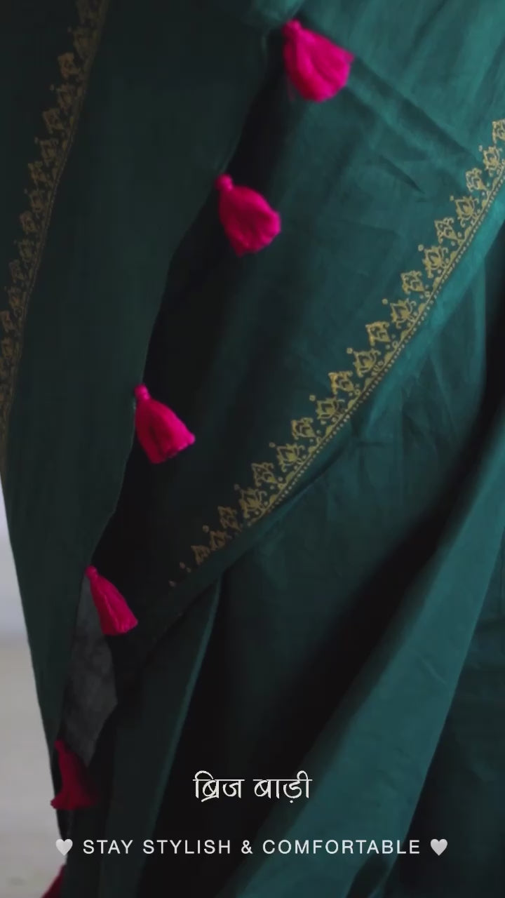 Deep green soft and light-weight cotton saree by Brij Bari, featuring a delicate hand block printed border in vibrant ochre yellow that contrasts beautifully with the rich green. The saree is finished with pink handmade thread tassels on the pallu, adding a touch of traditional charm. It comes with a free matching block printed blouse piece, completing the handcrafted look. Perfect for everyday wear, casual outings, or semi-formal occasions, with free shipping pan India.