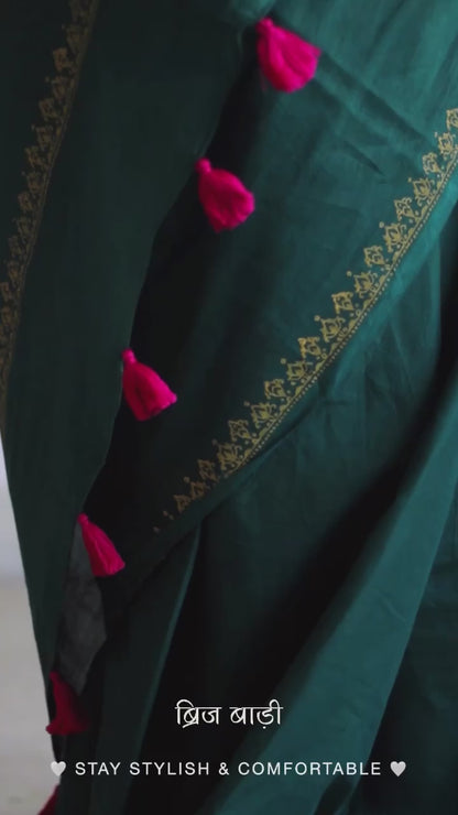 Deep green soft and light-weight cotton saree by Brij Bari, featuring a delicate hand block printed border in vibrant ochre yellow that contrasts beautifully with the rich green. The saree is finished with pink handmade thread tassels on the pallu, adding a touch of traditional charm. It comes with a free matching block printed blouse piece, completing the handcrafted look. Perfect for everyday wear, casual outings, or semi-formal occasions, with free shipping pan India.