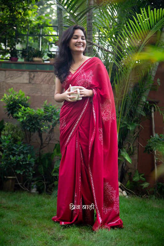 Deep red buttery soft and lightweight cotton saree by Brij Bari, hand block printed with delicate white rose motifs for a refined, artisanal touch. The pallu is finished with olive green handmade thread tassels that add a subtle contrast and handcrafted charm. Comes with a free coordinated block printed blouse piece. Designed for all-day comfort and understated elegance, this saree is a versatile pick for everyday wear or casual occasions. Free shipping pan India.