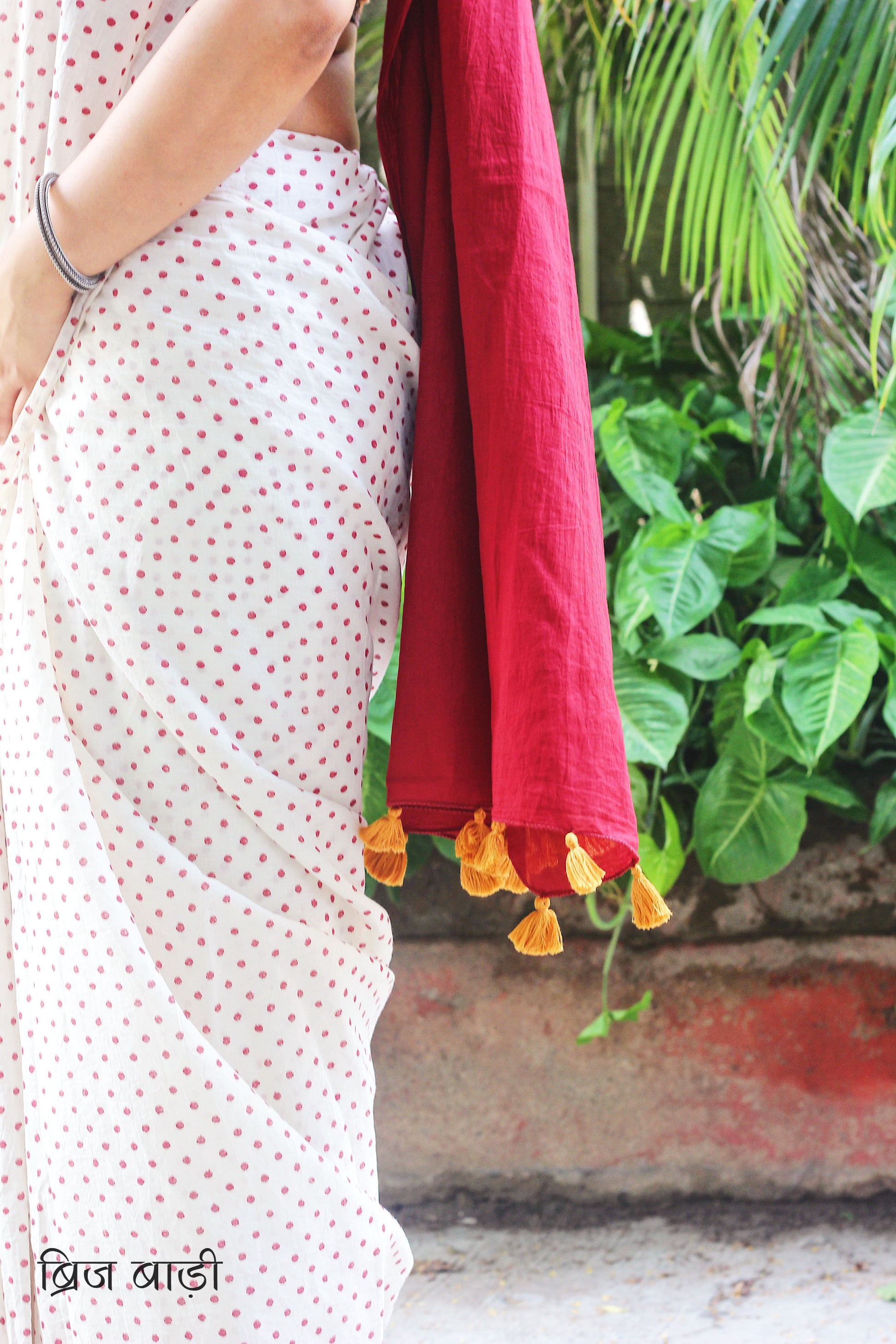 Off-white soft and light-weight cotton saree by Brij Bari, featuring deep red ombre pallu and small retro-style polka dot hand block prints. Accented with handcrafted mustard thread tassels on the pallu for a touch of artisanal charm. Includes a free matching hand block printed blouse piece. A timeless piece combining elegance, tradition, and comfort—crafted by Brij Bari’s skilled artisans.
