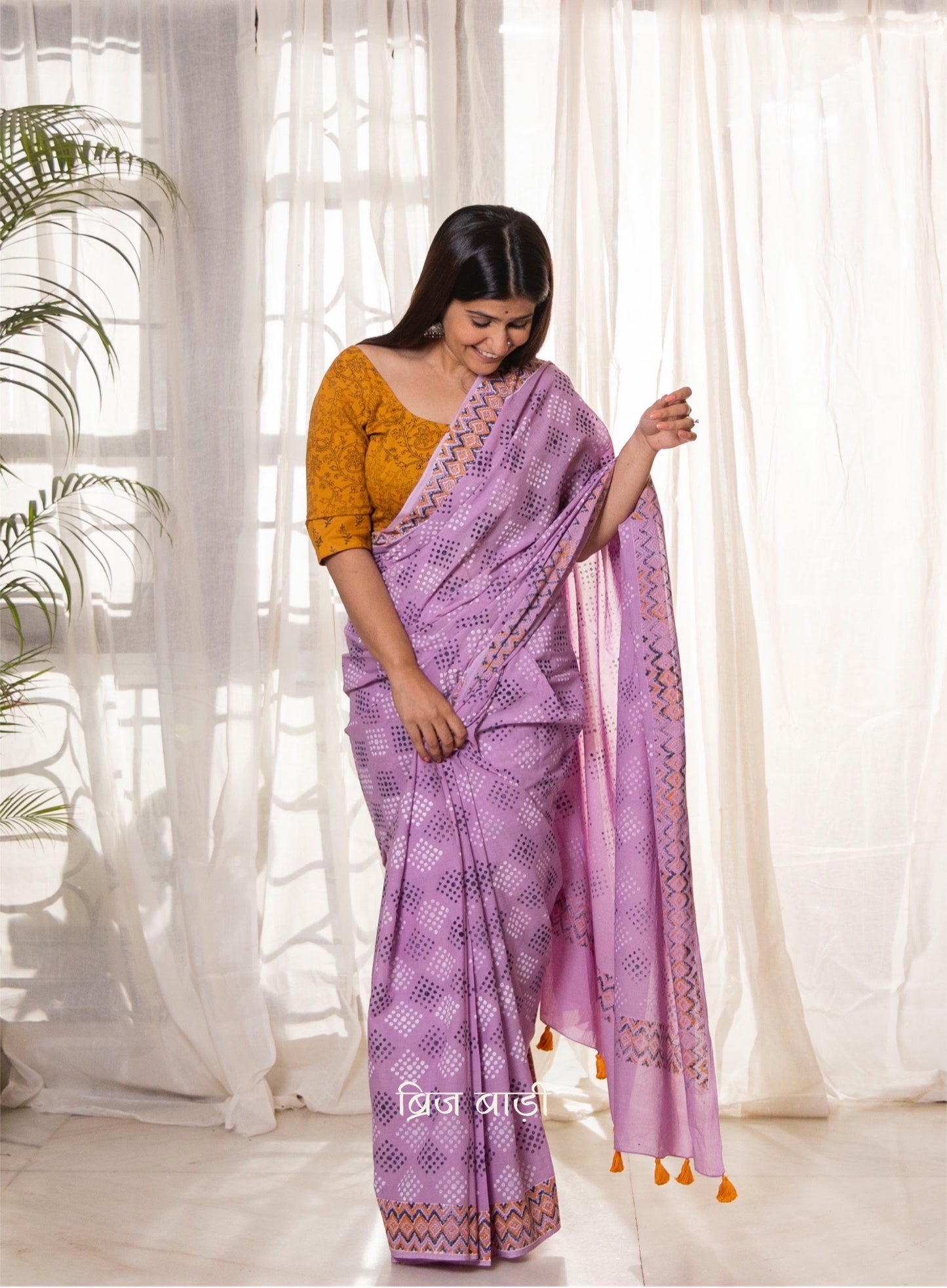 Lavender buttery soft and light-weight cotton saree by Brij Bari, featuring a playful polka dot pattern hand block printed in blue and yellow on the pallu. The saree is detailed with orange handmade thread tassels on the pallu, adding a pop of color. It comes with a free block printed blouse piece, making it a versatile option for everyday wear and casual occasions. Designed for all-day comfort and style, with free shipping available pan India.