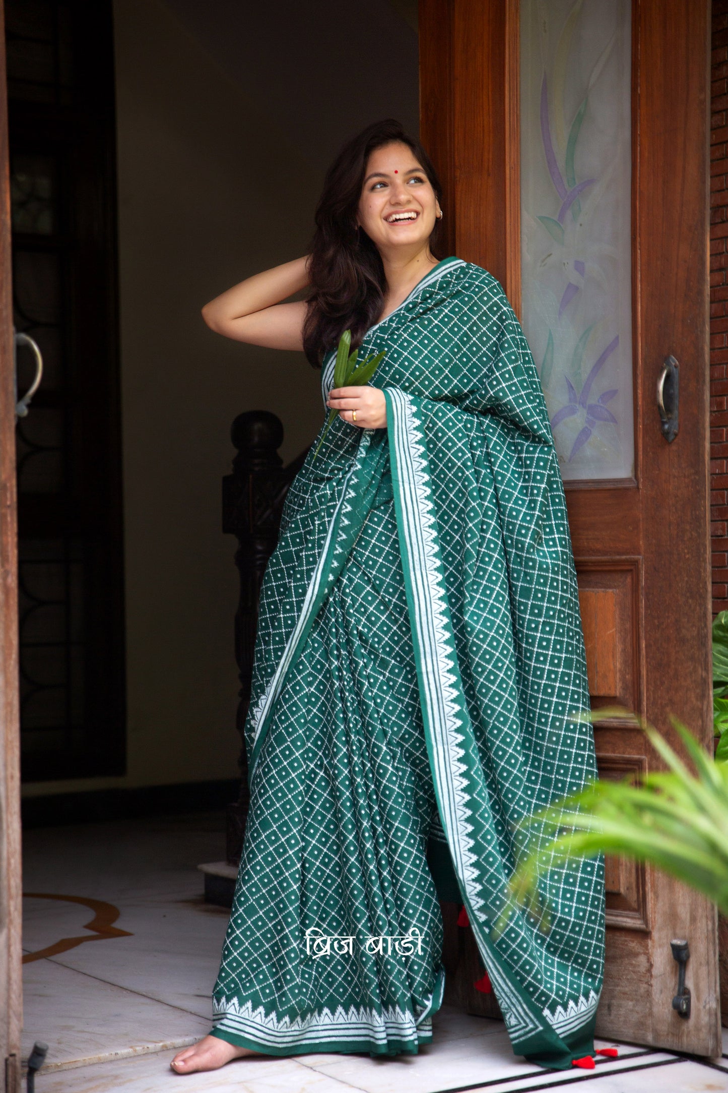 Dark green buttery soft and lightweight cotton saree by Brij Bari, hand block printed with an elegant white geometrical pattern. The pallu is accented with handcrafted red thread tassels, adding a vibrant artisanal detail. Includes a matching block printed blouse piece. Designed for all-day comfort and ease, this saree is a stylish and breathable option for everyday elegance. Free shipping pan India.