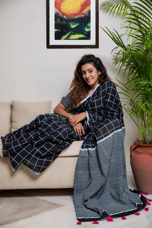 Black soft and light-weight cotton saree by Brij Bari, hand block printed in white with a classic check pattern on the body and a striking geometrical pattern on the pallu. Detailed with pink handmade thread tassels that add a charming, handcrafted touch. This saree comes with a free matching block printed blouse piece, offering a complete, artisanal look. A versatile, breathable saree perfect for everyday wear, casual outings, or semi-formal occasions, with free shipping pan India. Available in 2 colours