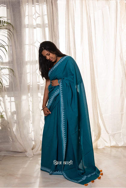 Teal blue soft and light-weight cotton saree by Brij Bari, designed for all-day comfort and effortless elegance. Detailed with a delicate hand block printed border in crisp white, creating a refined contrast. The saree is finished with orange handmade thread tassels on the pallu, adding a subtle pop of colour. Comes with a free matching block printed blouse piece. Ideal for everyday wear, office, casual outings, or light festive occasions, with free shipping pan India.