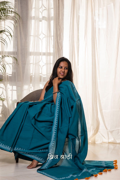 Teal blue soft and light-weight cotton saree by Brij Bari, designed for all-day comfort and effortless elegance. Detailed with a delicate hand block printed border in crisp white, creating a refined contrast. The saree is finished with orange handmade thread tassels on the pallu, adding a subtle pop of colour. Comes with a free matching block printed blouse piece. Ideal for everyday wear, office, casual outings, or light festive occasions, with free shipping pan India.