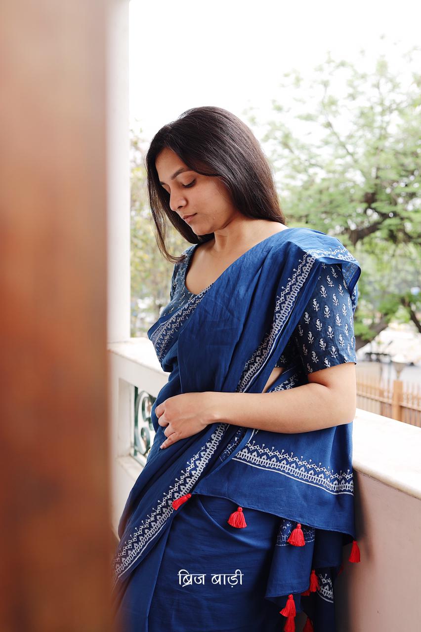 Blue soft and light-weight cotton saree by Brij Bari hand block printed with an elegant white border pattern that lends a timeless, understated charm. Made from breathable cotton fabric, this saree ensures day-long comfort making it perfect for everyday wear, work or casual outings. The pallu is adorned with bright red handmade thread tassels adding a vibrant handcrafted accent to the look. It comes with a free hand block printed blouse piece.  Free shipping pan India.
 