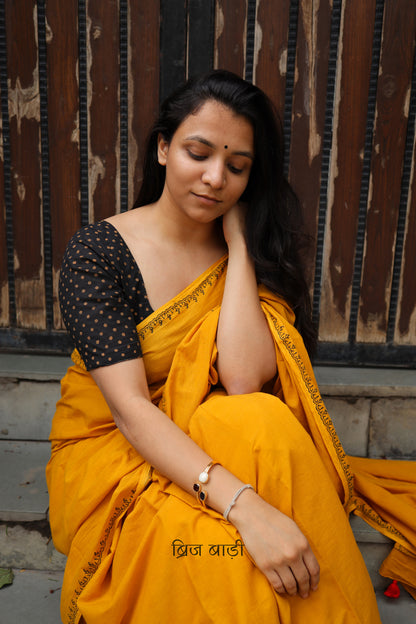 Yellow soft and light-weight cotton saree by Brij Bari hand block printed with a simple and elegant border pattern in classic black for a clean, timeless look. Crafted from breathable this saree is perfect for everyday wear easy to drape, comfortable for warm weather and effortlessly stylish. The pallu is adorned with delicate light green handmade thread tassels adding a fresh handcrafted touch to the design. Paired with a free hand block printed blouse piece carefully coordinated to match the saree